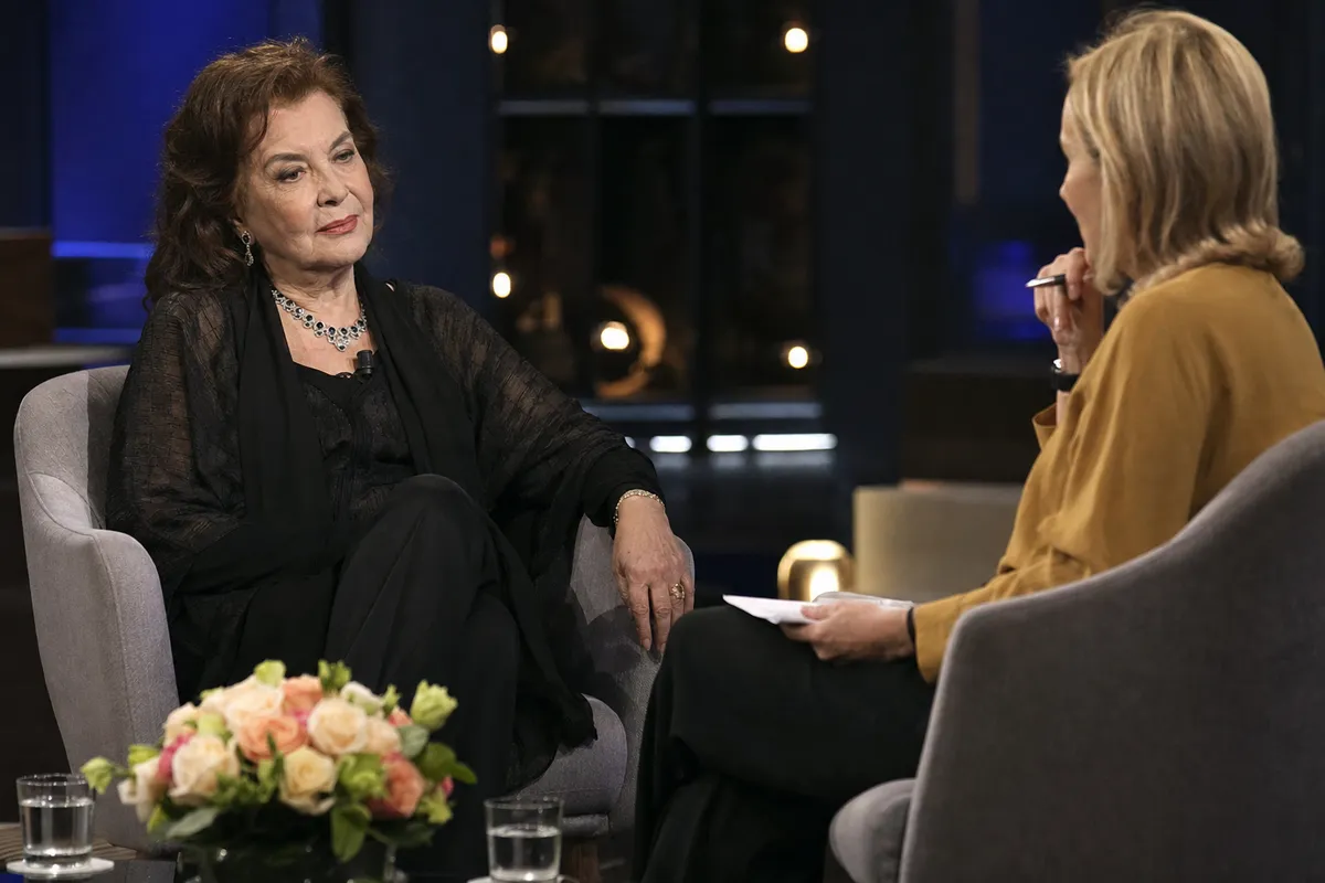 Intervista in studio TV: donna anziana elegante in poltrona parla con una conduttrice, bouquet di rose sul tavolino