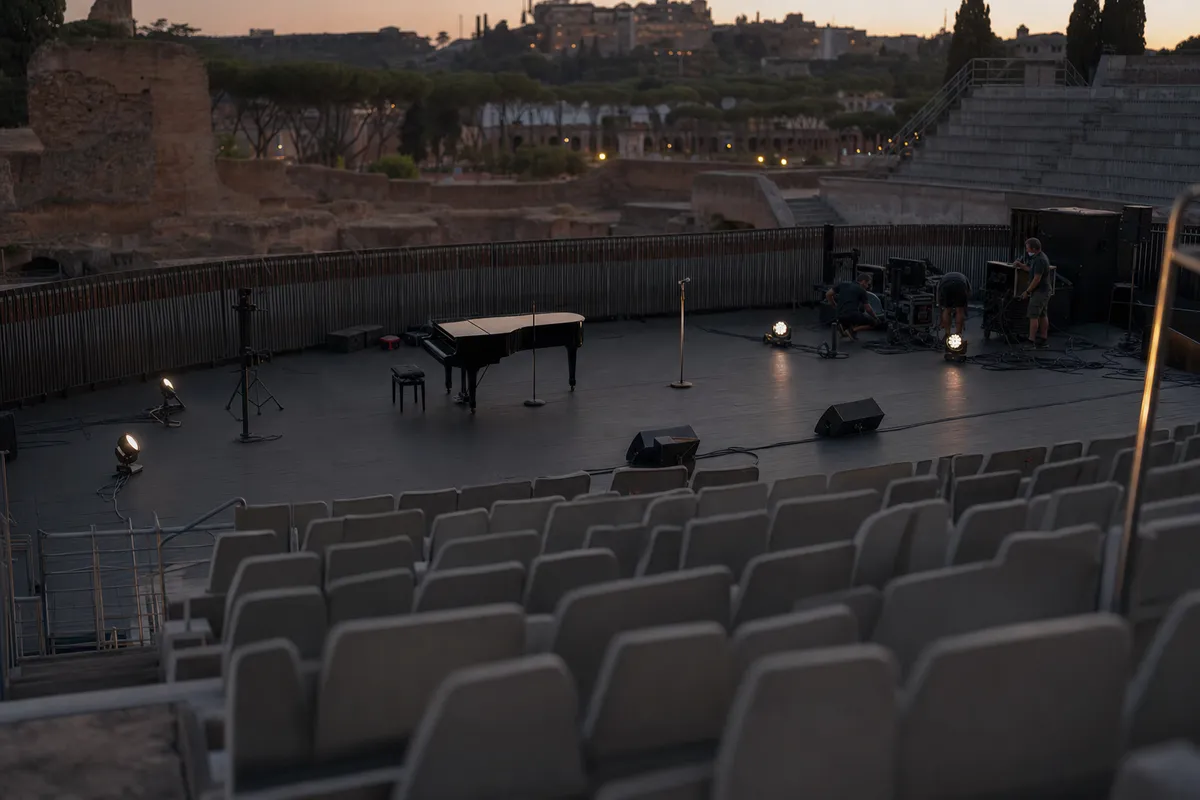 Palco vuoto in un anfiteatro all’aperto al tramonto, con pianoforte a coda, microfoni e tecnici al lavoro