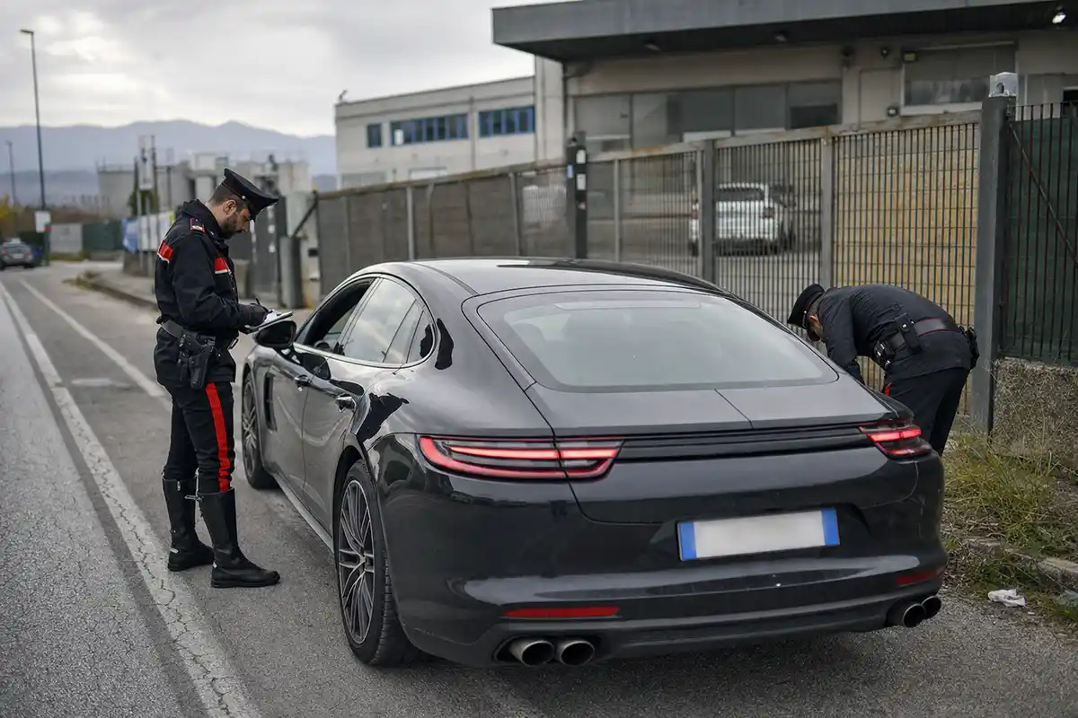 Due carabinieri controllano un’auto di lusso scura ferma a bordo strada vicino a un’area industriale