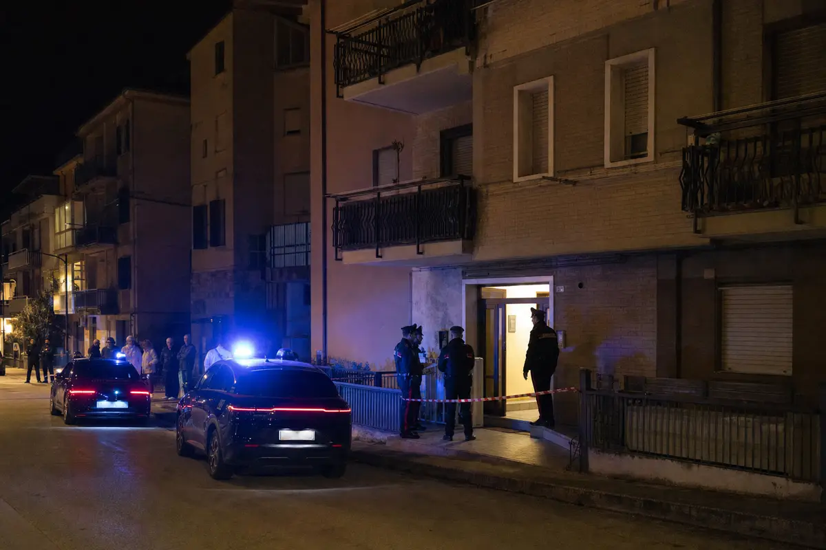 Carabinieri e auto di servizio con luci blu davanti a un condominio di notte, ingresso transennato