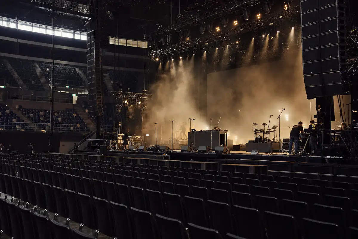 Palco in un’arena vuota durante il soundcheck, con luci calde, fumo scenico, microfoni e postazione DJ