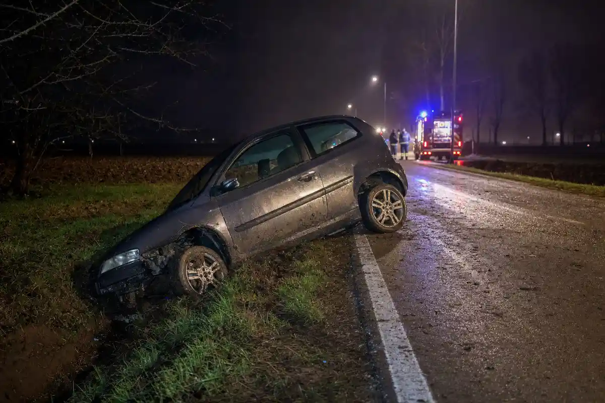 Auto finita in un fosso lungo una strada di campagna bagnata di notte, con luci di emergenza sullo sfondo
