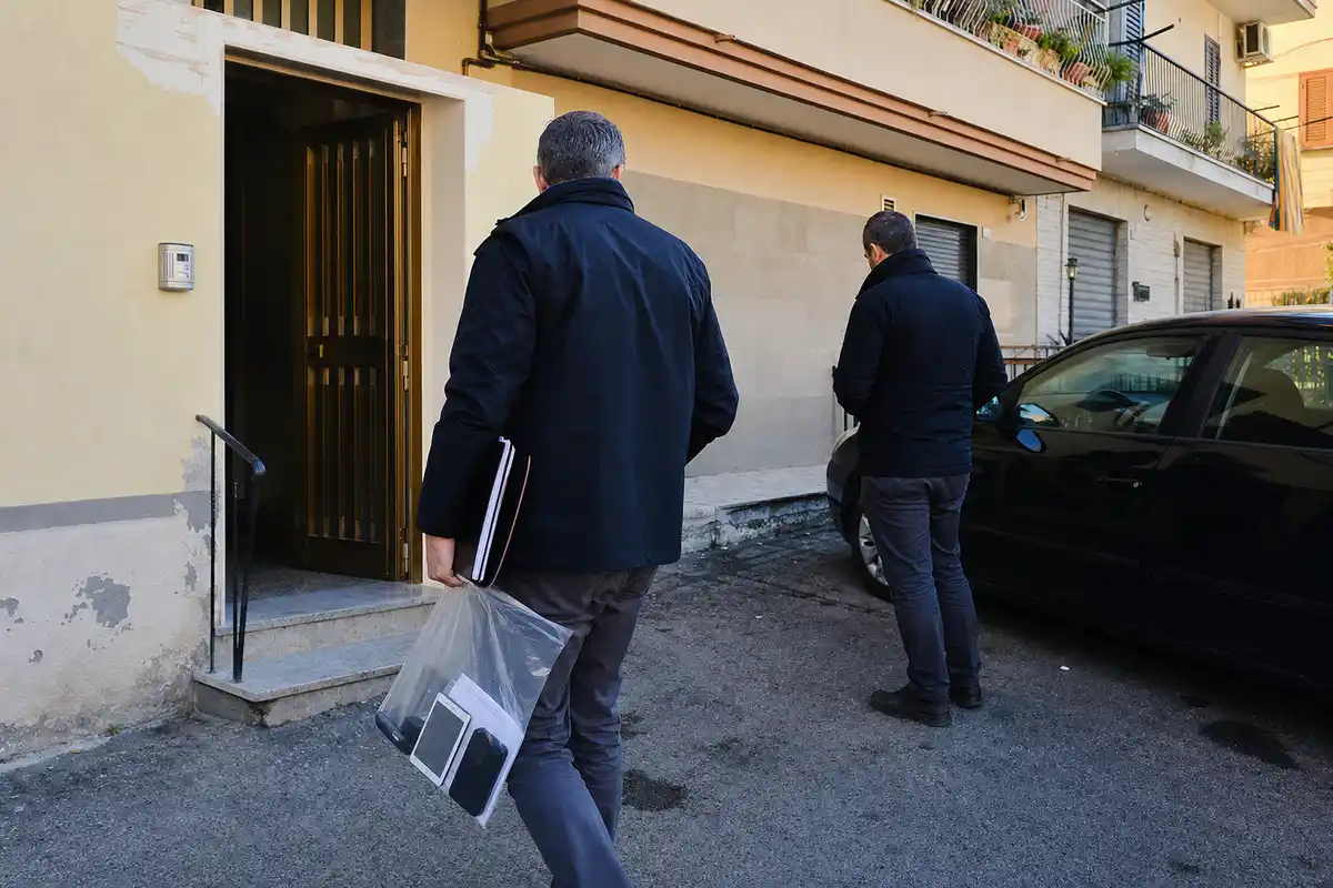 Due agenti davanti all’ingresso di un palazzo, uno con busta prove e fascicoli accanto a un’auto scura