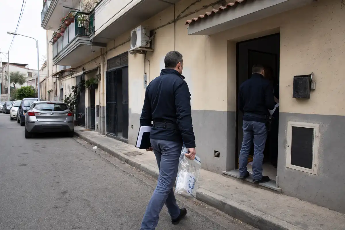 Due agenti in divisa scura fuori da un portone: uno entra nell’abitazione, l’altro tiene documenti e una busta sigillata
