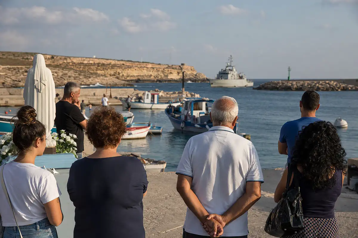 Residenti osservano il mare nel porto, con statua religiosa e barche da pesca; sullo sfondo una nave di pattugliamento
