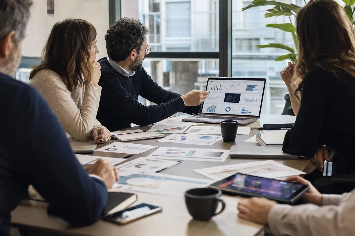 Riunione in ufficio con professionisti attorno a un tavolo, report cartacei e laptop con grafici e dashboard