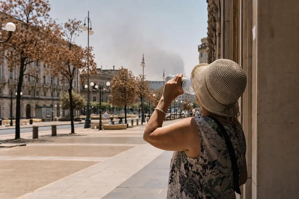 Donna con cappello di paglia beve acqua all’ombra su un viale cittadino assolato e quasi deserto, con foschia sullo sfondo