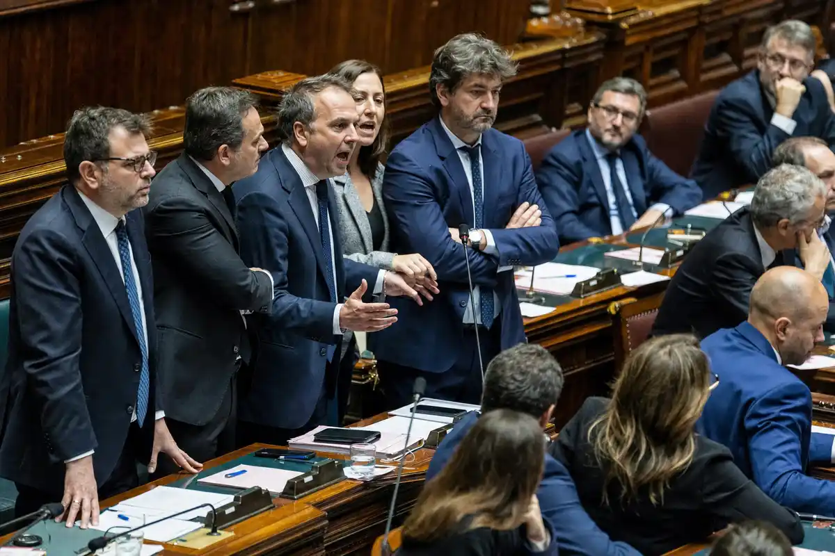 Deputati in abiti formali discutono animatamente in Aula tra banchi in legno, microfoni e documenti