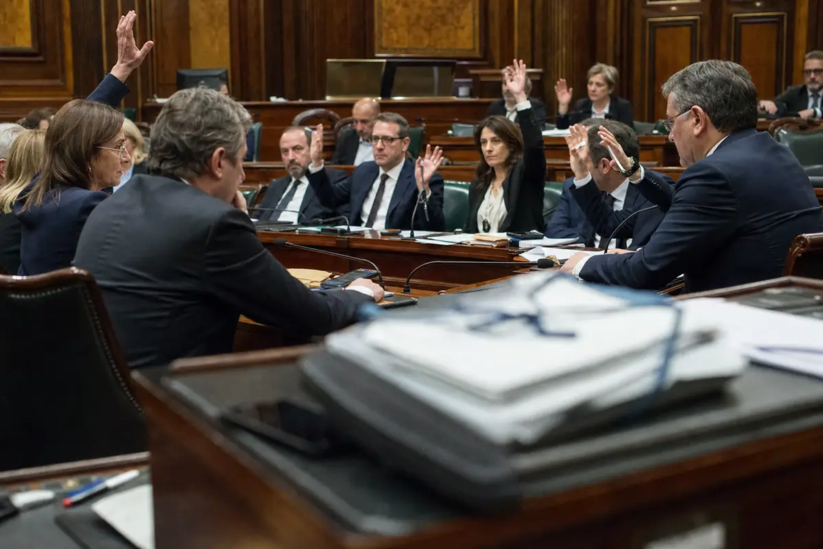 Parlamentari in una sala istituzionale alzano la mano durante una votazione, con fascicoli e documenti in primo piano