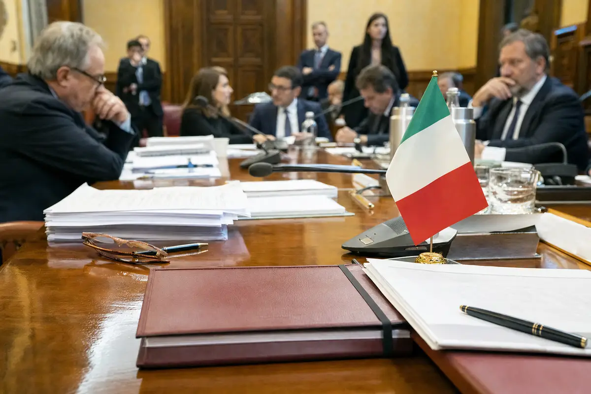 Tavolo di commissione parlamentare con documenti, cartelline e bandierina italiana, deputati sfocati sullo sfondo