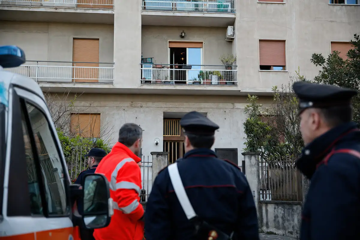 Facciata di palazzo con balconi e finestra aperta, con carabinieri e soccorritori in primo piano vicino a un’ambulanza