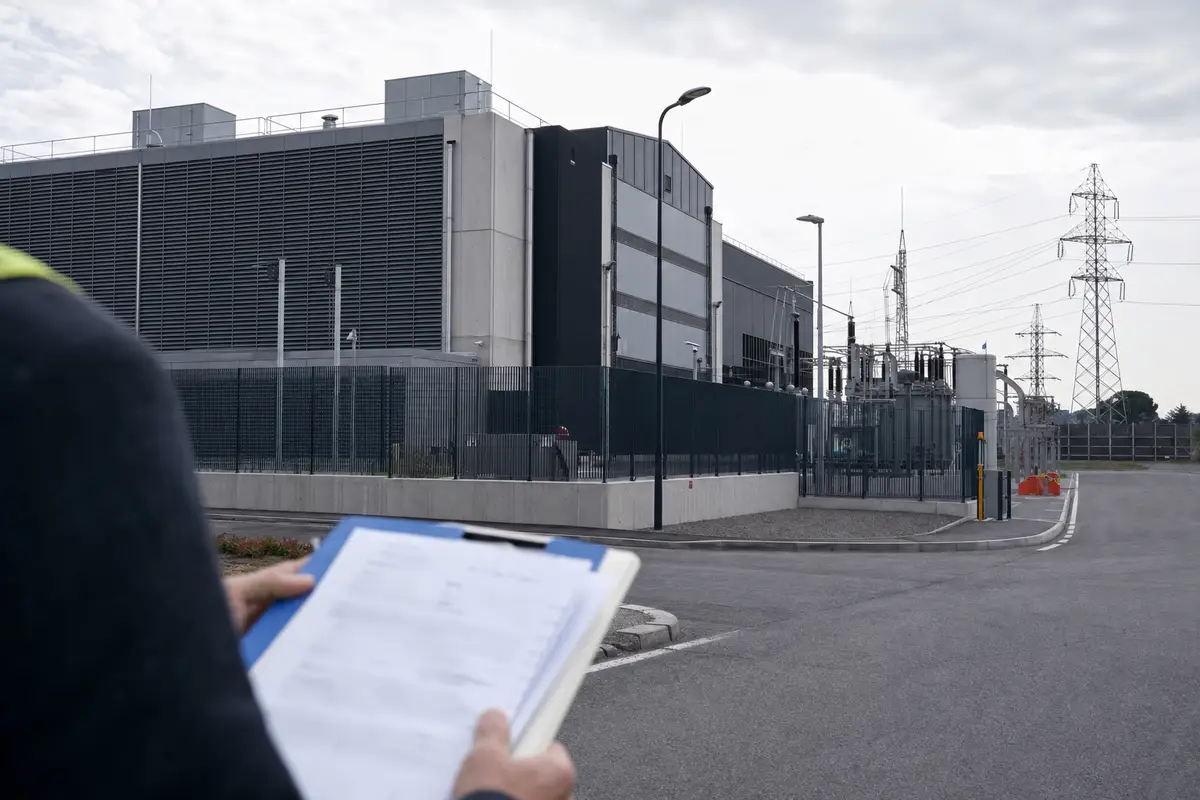 Tecnico con cartellina in mano davanti a un moderno data center recintato con sottostazione elettrica e tralicci sullo sfondo