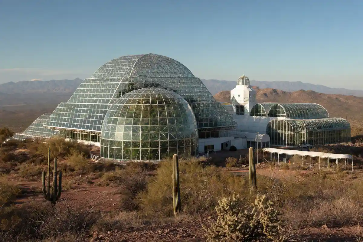 Struttura di ricerca in vetro con grandi cupole nel deserto, tra cactus e colline, con vegetazione visibile all’interno