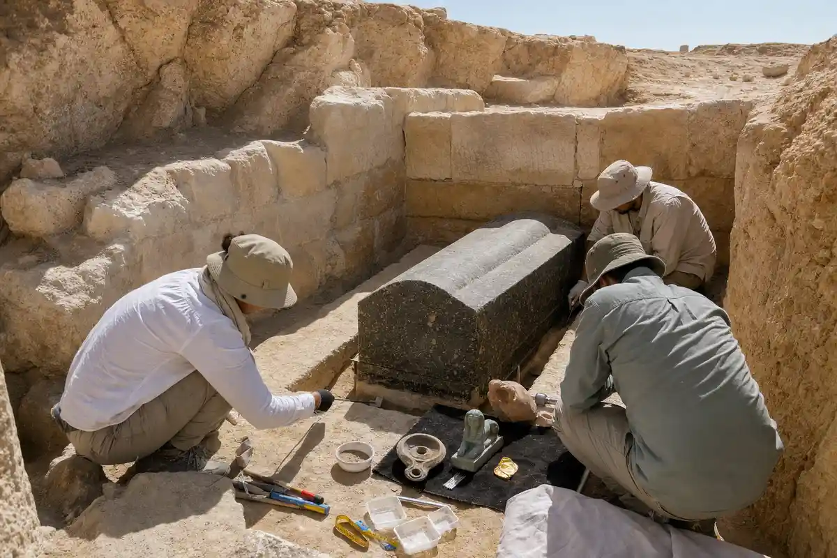 Archeologi al lavoro in una tomba scavata nella roccia con un grande sarcofago di granito e reperti sul telo