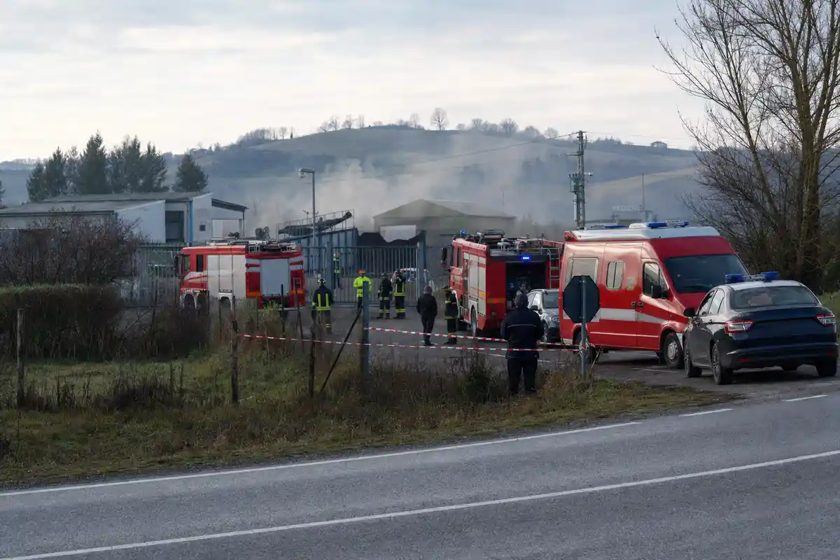 Mezzi di soccorso e vigili del fuoco davanti al cancello di un impianto industriale, con nastro di sicurezza e lieve fumo
