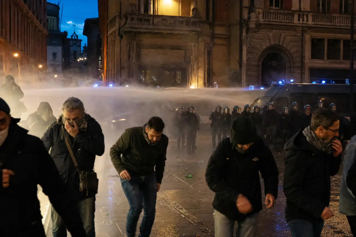 Manifestanti si allontanano tra fumo e strada bagnata, con polizia in tenuta antisommossa e blindati sullo sfondo al