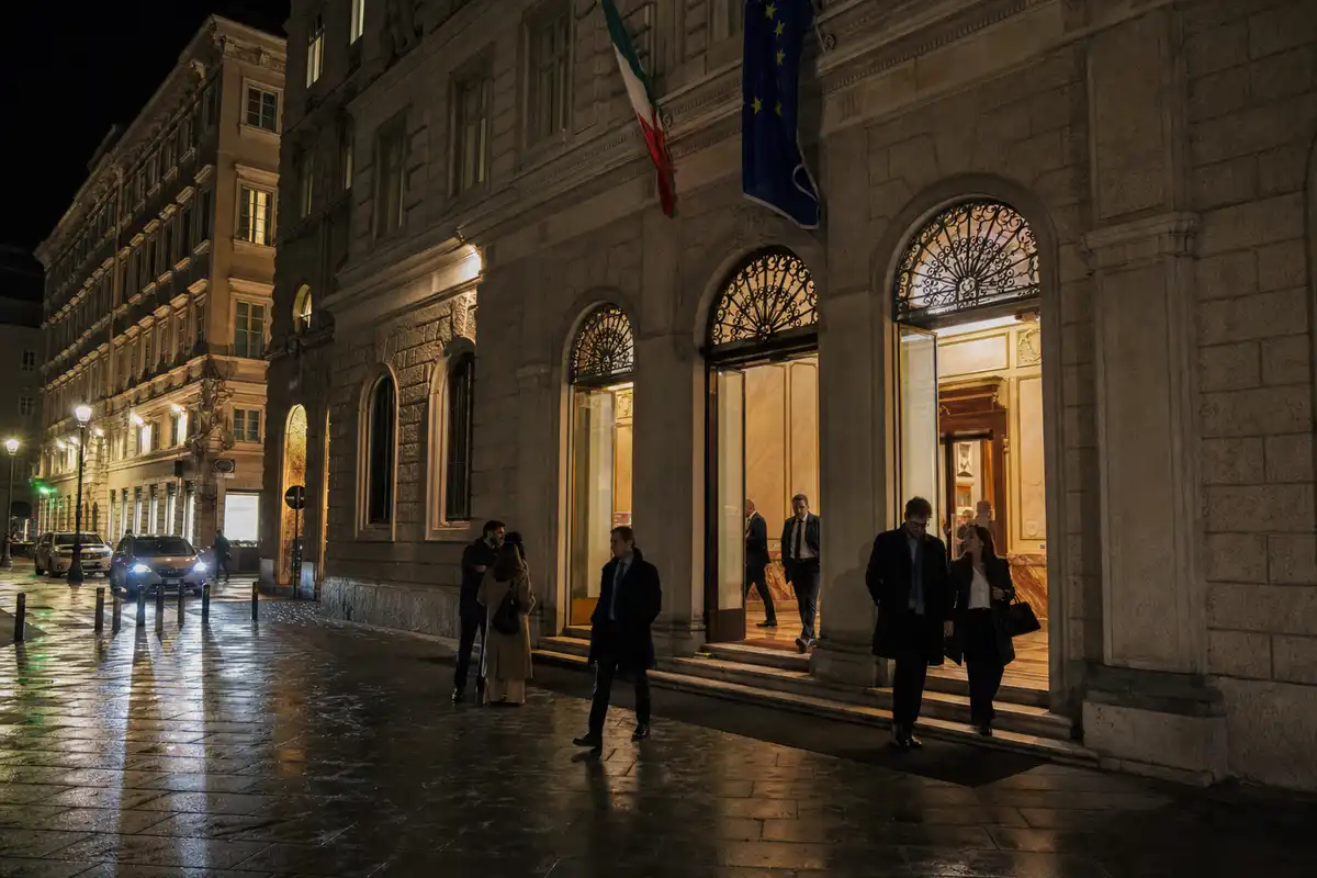 Ingresso illuminato di un palazzo istituzionale di notte, con bandiere italiana ed europea e persone che escono sul