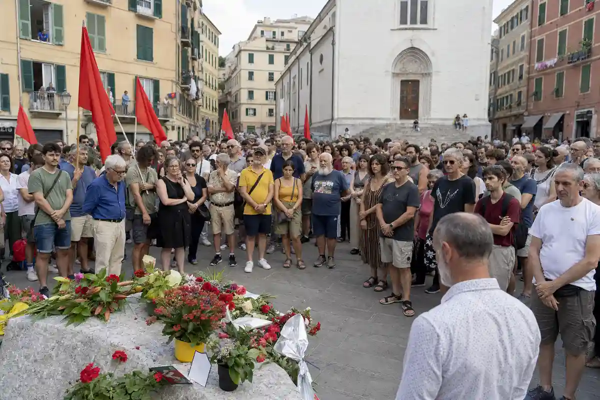 Folla in piazza davanti a un cippo con mazzi di fiori, con bandiere rosse e una chiesa sullo sfondo