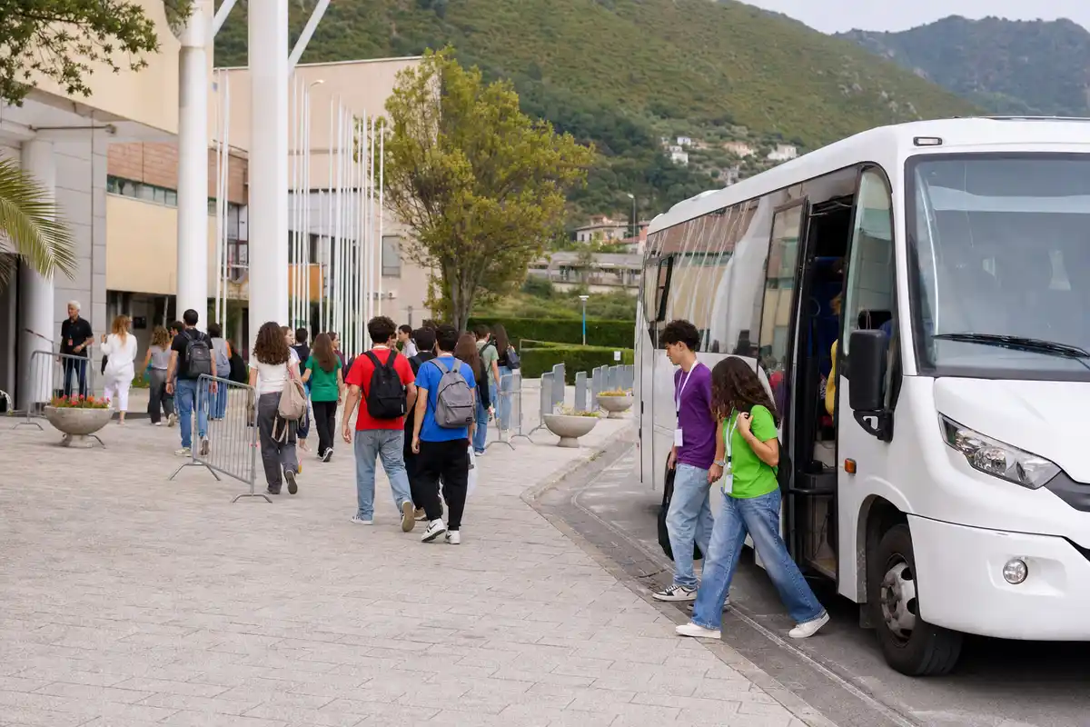 Navetta bianca con giovani partecipanti che scendono e camminano verso l’ingresso di un festival, con colline sullo sfondo