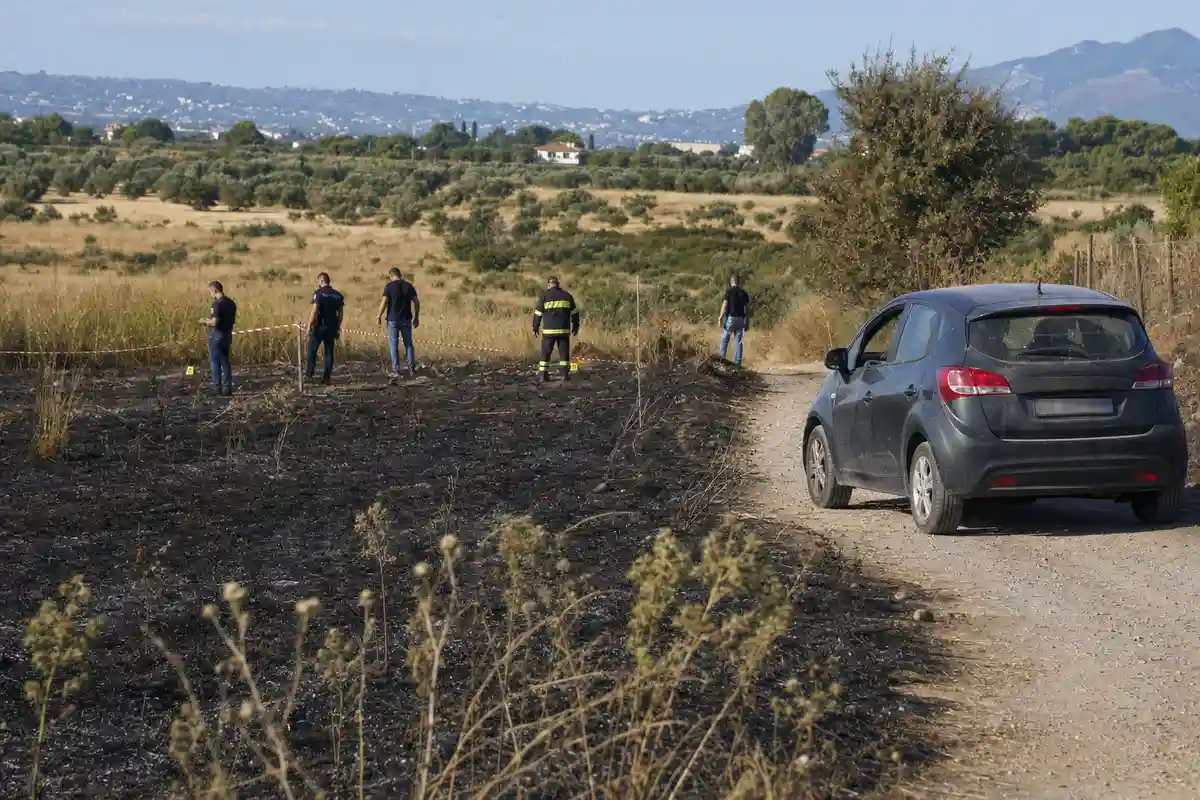 Auto compatta su strada sterrata accanto a campo bruciato con investigatori e nastro segnaletico
