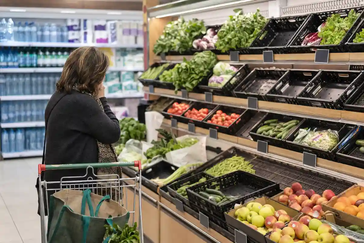 Cliente al supermercato guarda i prezzi nel reparto ortofrutta, con carrello e borsa riutilizzabile