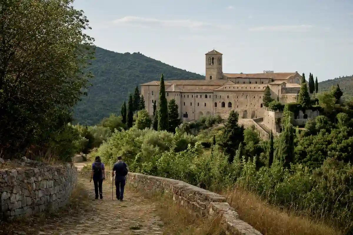 Due pellegrini di spalle camminano su un sentiero in pietra verso un’abbazia tra colline verdi e cipressi
