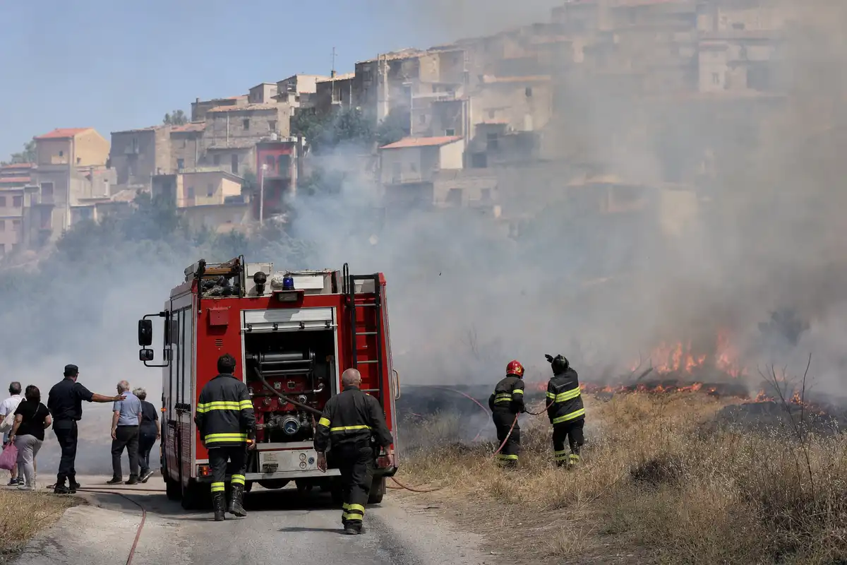 Camion dei vigili del fuoco e pompieri con tubo mentre un incendio di sterpaglie minaccia un paese su una collina