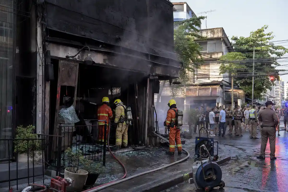 Vigili del fuoco davanti all’ingresso annerito di un pub dopo un incendio, con fumo, tubi e folla sullo sfondo