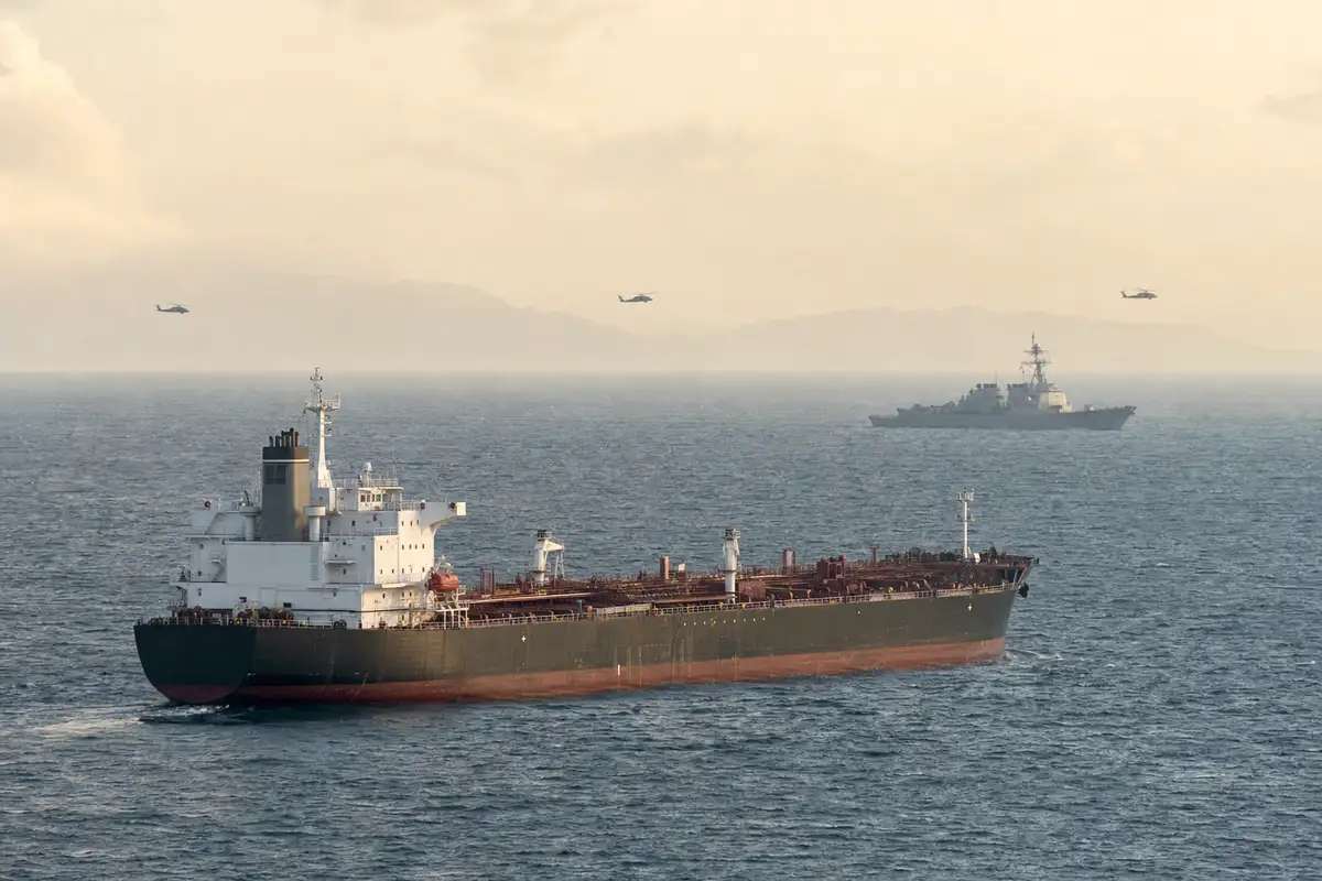 Petroliera in mare con nave militare sullo sfondo e elicotteri in volo in un cielo velato