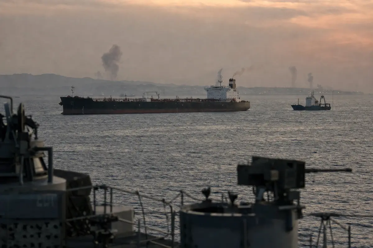 Petroliera e nave cargo in mare al tramonto, con colonne di fumo lontane e sagoma di nave militare in primo piano