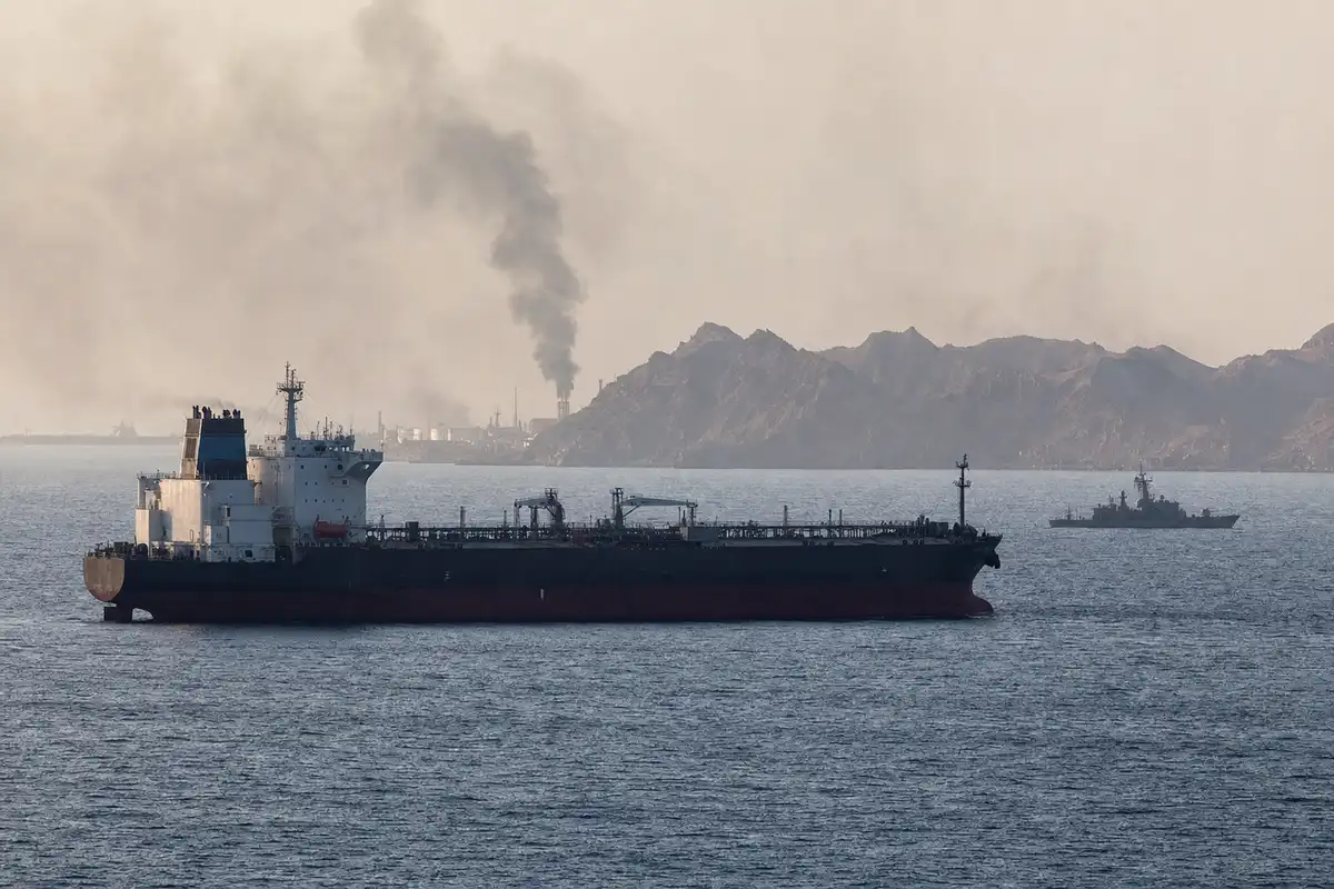 Petroliera in mare con nave militare all’orizzonte e colonna di fumo vicino alla costa rocciosa