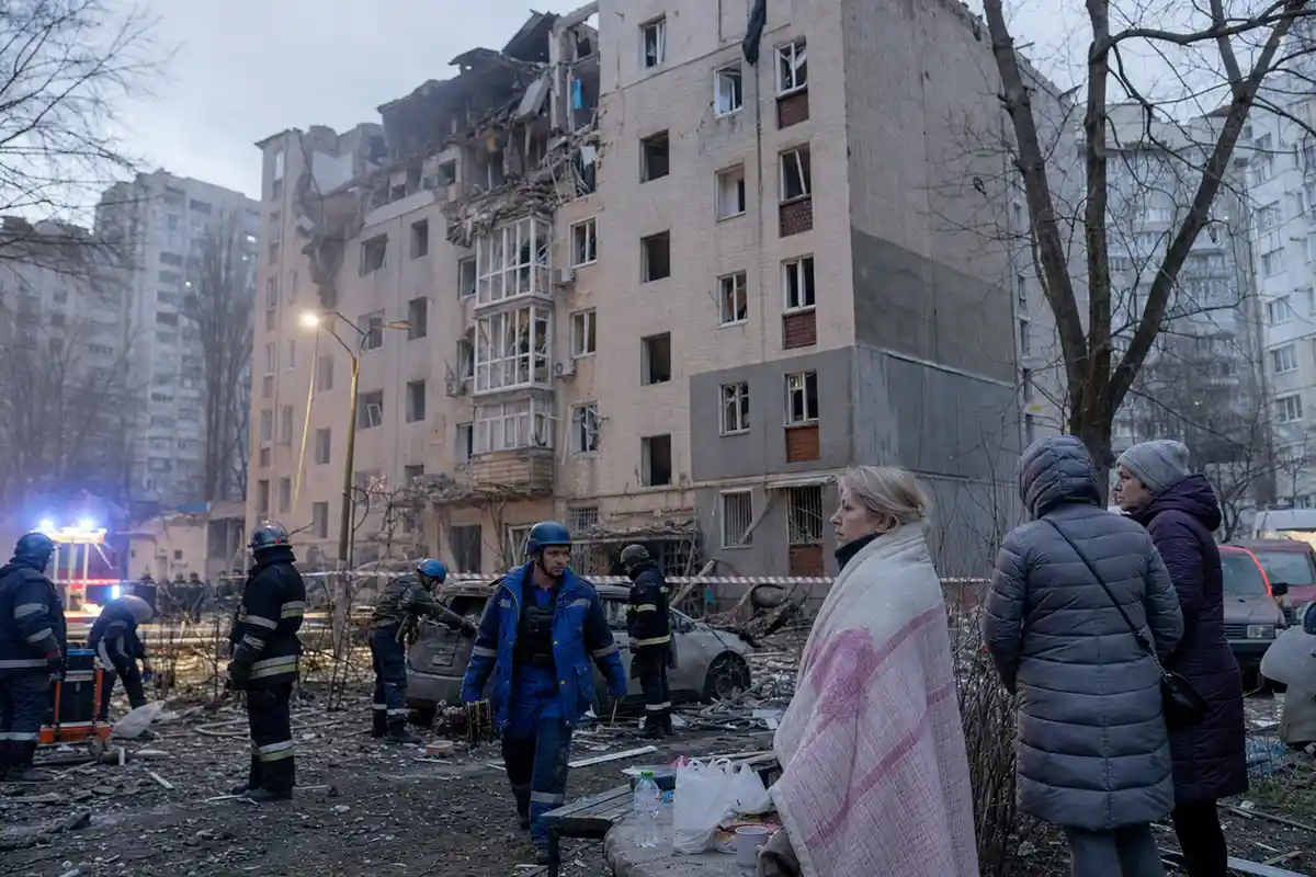 Soccorritori e residenti davanti a un palazzo residenziale danneggiato, con macerie e auto colpite all’alba
