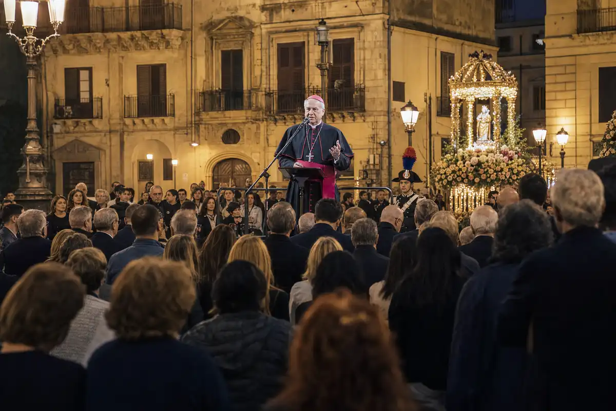 Vescovo sul palco parla al microfono davanti a una folla in piazza di sera, con luminarie e fercolo religioso