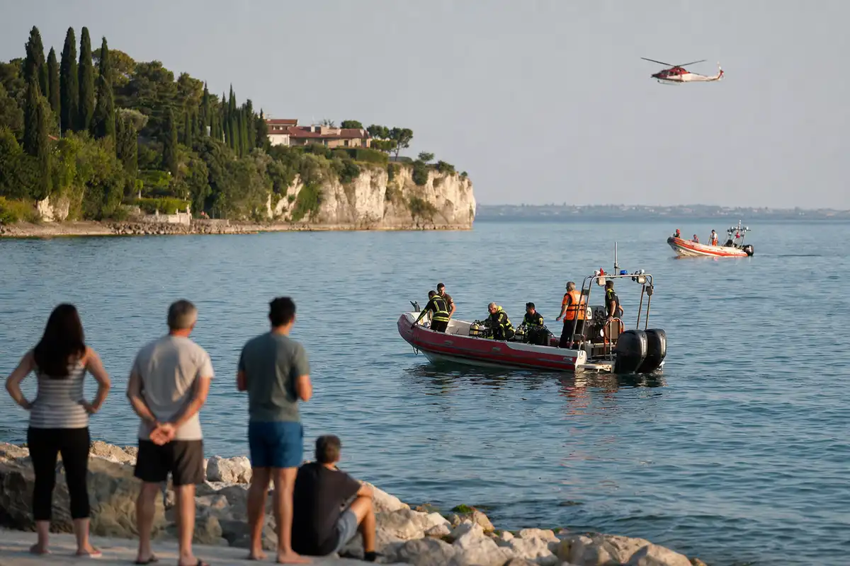 Barche di soccorso in operazione sul lago con elicottero in volo e bagnanti che osservano dalla riva rocciosa