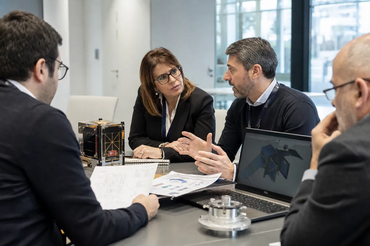 Professionisti in riunione attorno a un tavolo con documenti tecnici, laptop e un piccolo componente satellitare