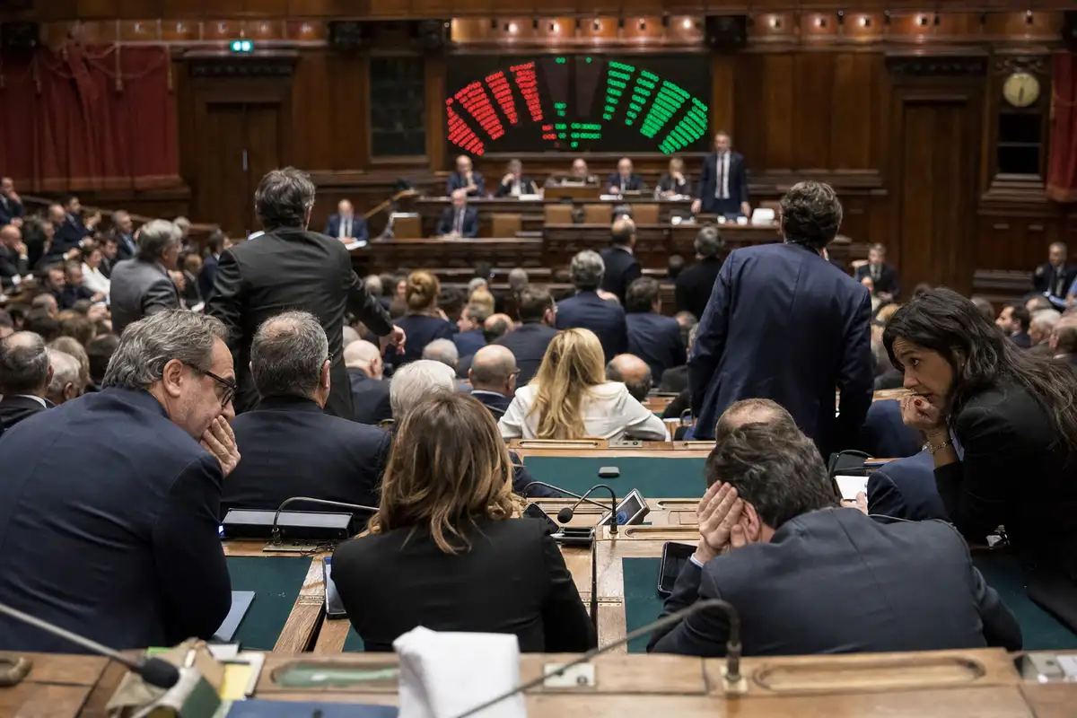 Aula parlamentare con deputati ai banchi e tabellone elettronico di voto con luci rosse e verdi sullo sfondo