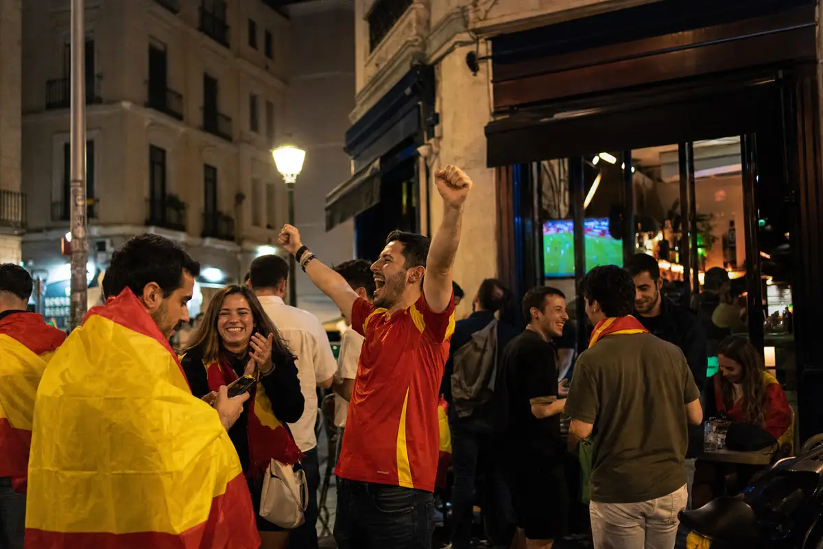 Tifosi con bandiere spagnole festeggiano fuori da un bar di notte, con una partita in TV visibile dalla vetrina