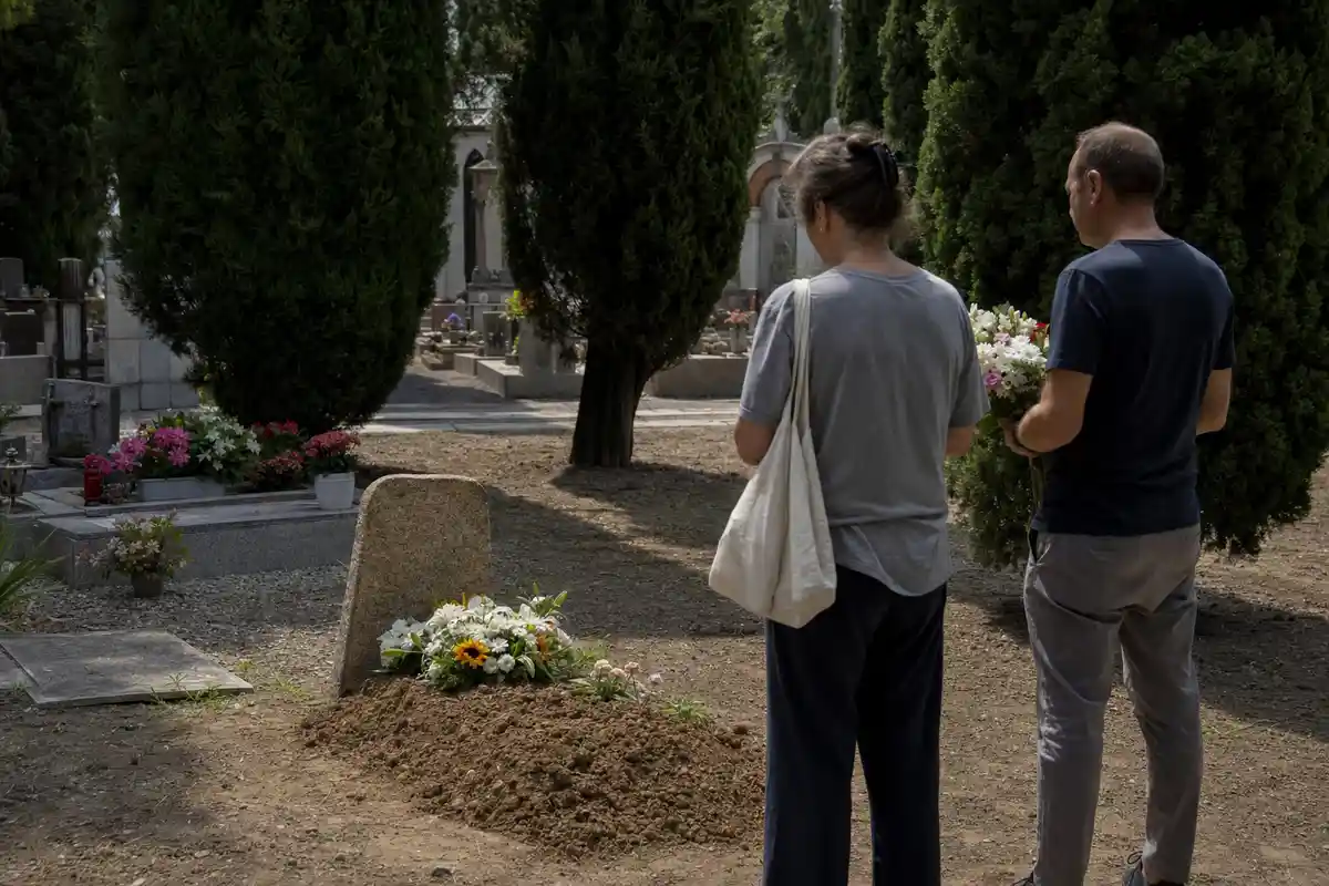 Due persone di spalle davanti a una tomba semplice con fiori in un cimitero, tra cipressi