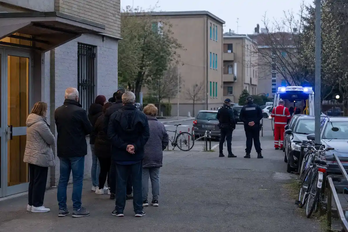 Residenti davanti a un palazzo osservano polizia e soccorritori vicino a un’ambulanza con luci accese