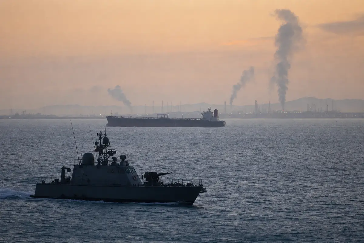 Motovedetta militare in primo piano e petroliera al largo all’alba, con colonne di fumo vicino a un porto industriale