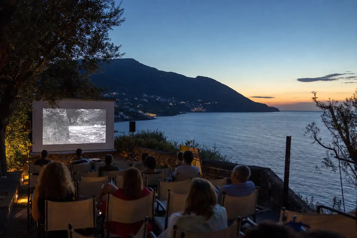 Proiezione all’aperto al tramonto su una terrazza affacciata sul mare, con schermo e pubblico seduto