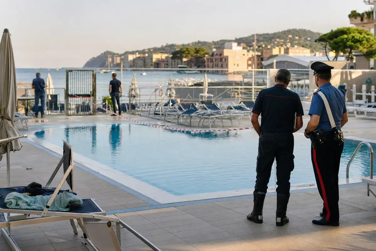 Piscina all’aperto transennata con due uomini in divisa a bordo vasca, lettini vuoti e mare sullo sfondo