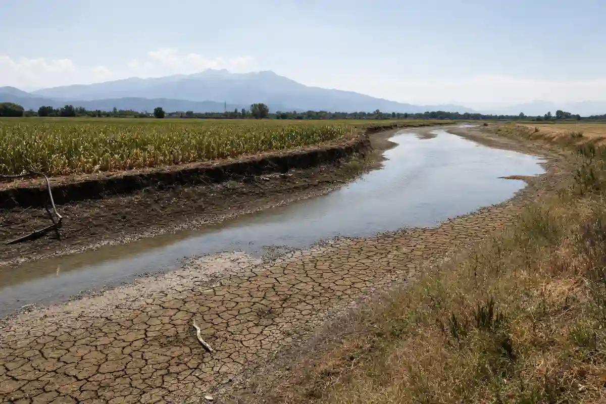 Letto di fiume in secca con fango screpolato, esile corso d’acqua e campi coltivati sullo sfondo montano
