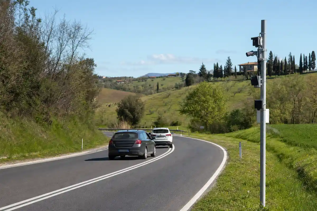 Strada extraurbana con doppia linea continua, due auto in curva e palo con telecamere di controllo a bordo carreggiata