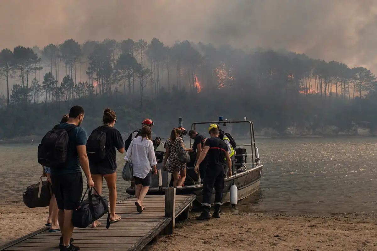 Evacuati con borse e zaini salgono su una barca di soccorso mentre un incendio boschivo brucia tra i pini