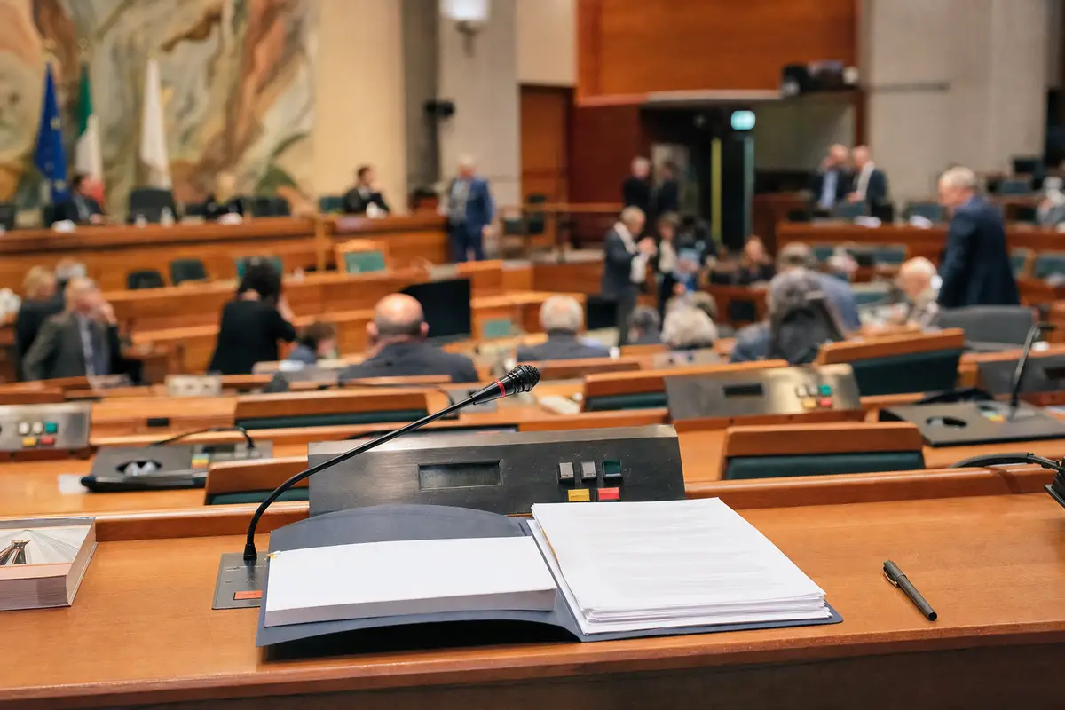 Banco in aula con microfono e fascicoli, sullo sfondo consiglieri sfocati in una sala assembleare