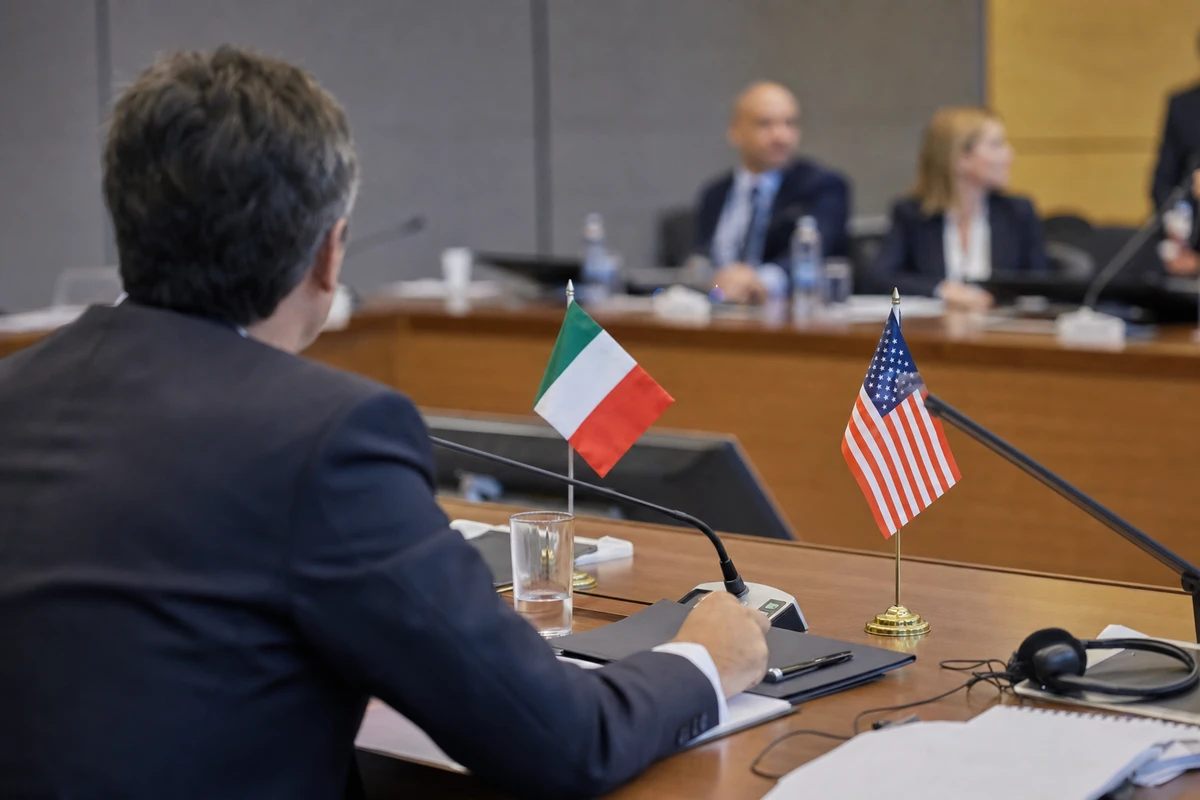 Delegato in giacca a un tavolo di riunione con bandierine di Italia e Stati Uniti e microfoni