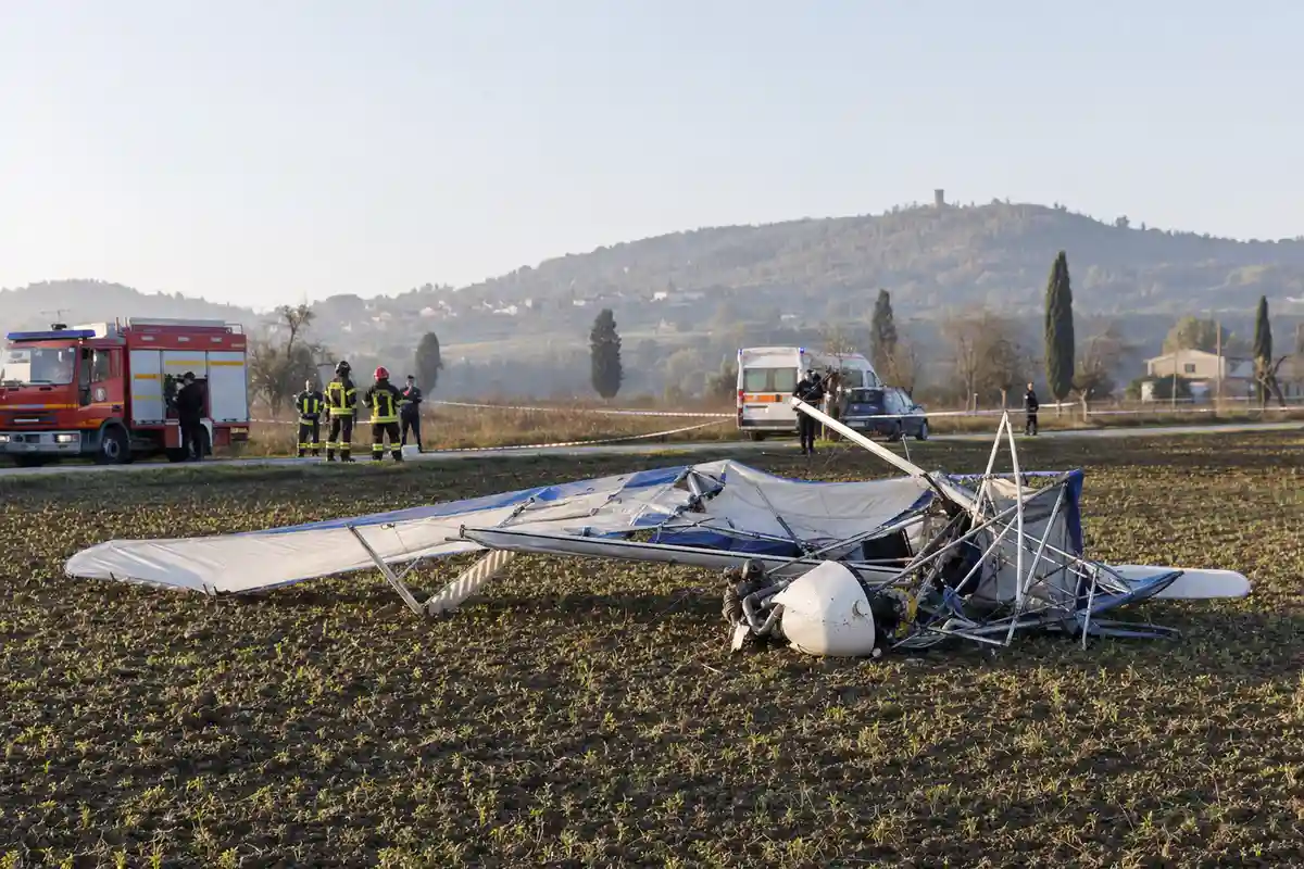 Rottami di un ultraleggero in un campo, con vigili del fuoco e ambulanza sullo sfondo dietro il nastro