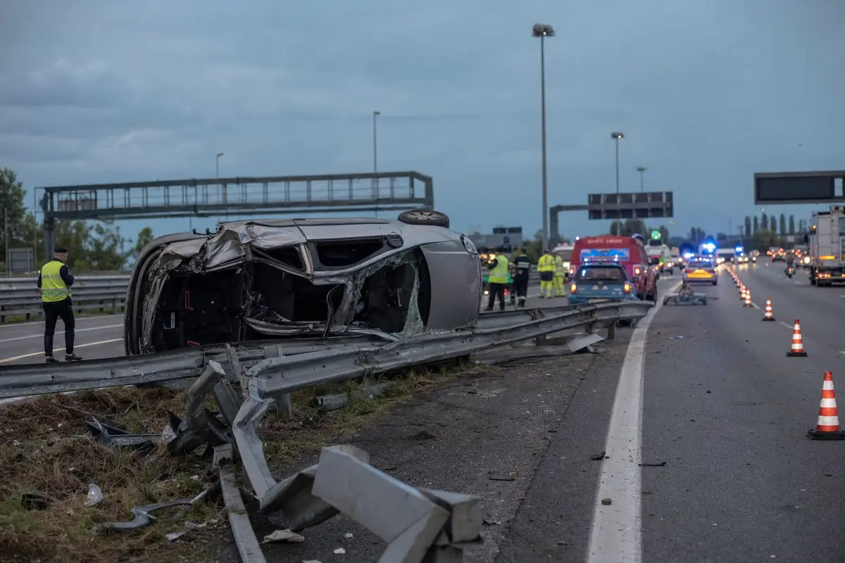 Minivan ribaltato contro il guardrail su autostrada, con coni e mezzi di soccorso sullo sfondo al crepuscolo