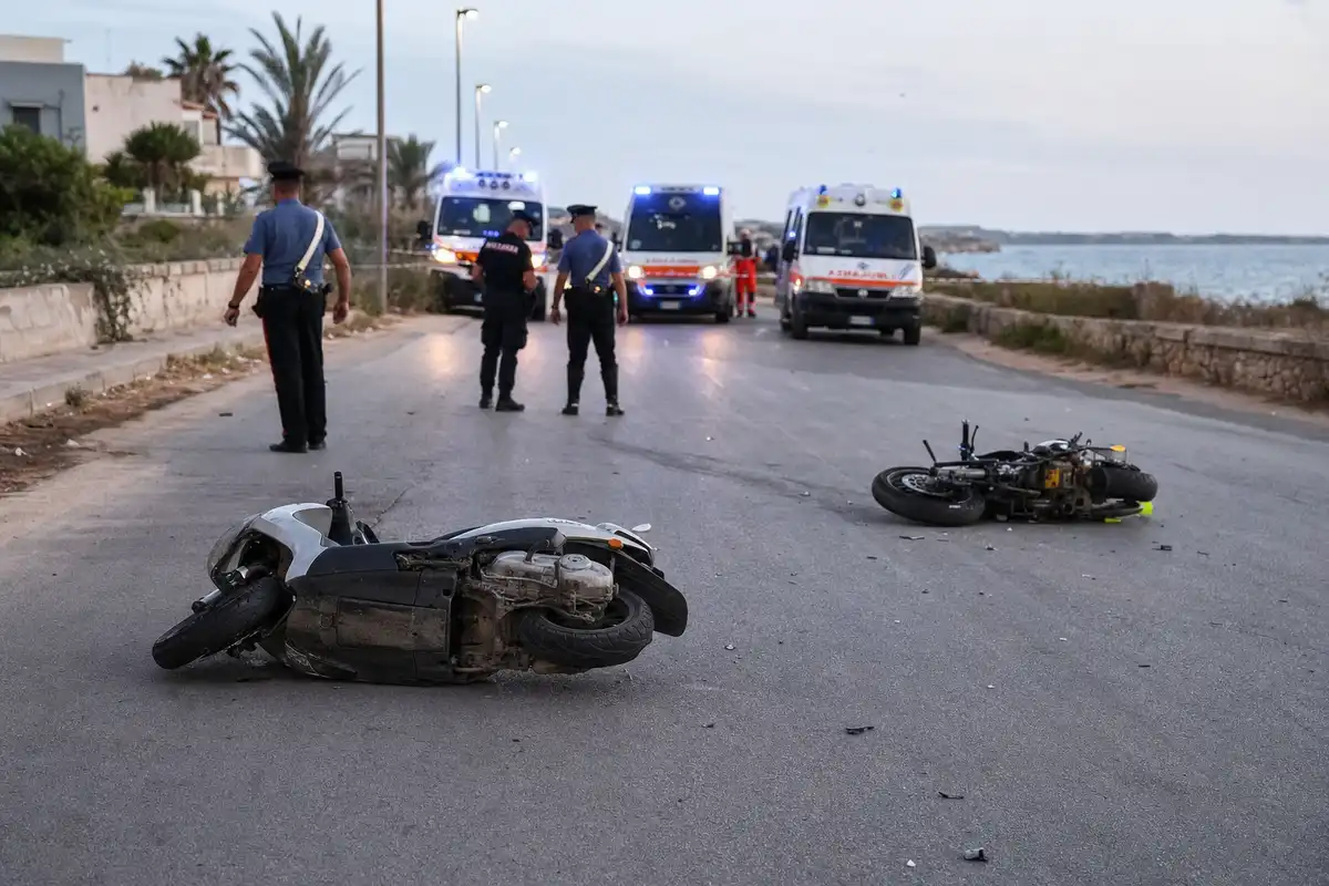 Scooter e moto danneggiati sull’asfalto con carabinieri e ambulanze sullo sfondo su strada costiera al tramonto